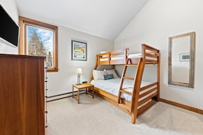 A bedroom with a bunk bed, a bedside table with a lamp, a framed picture on the wall, and a window with a view of trees.