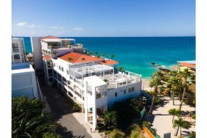 A coastal hotel with multiple balconies overlooking the sea, a beach area with palm trees, and a clear view of the ocean. - Maho Beach Modern 1-Bedroom, Ocean Level (Maho)