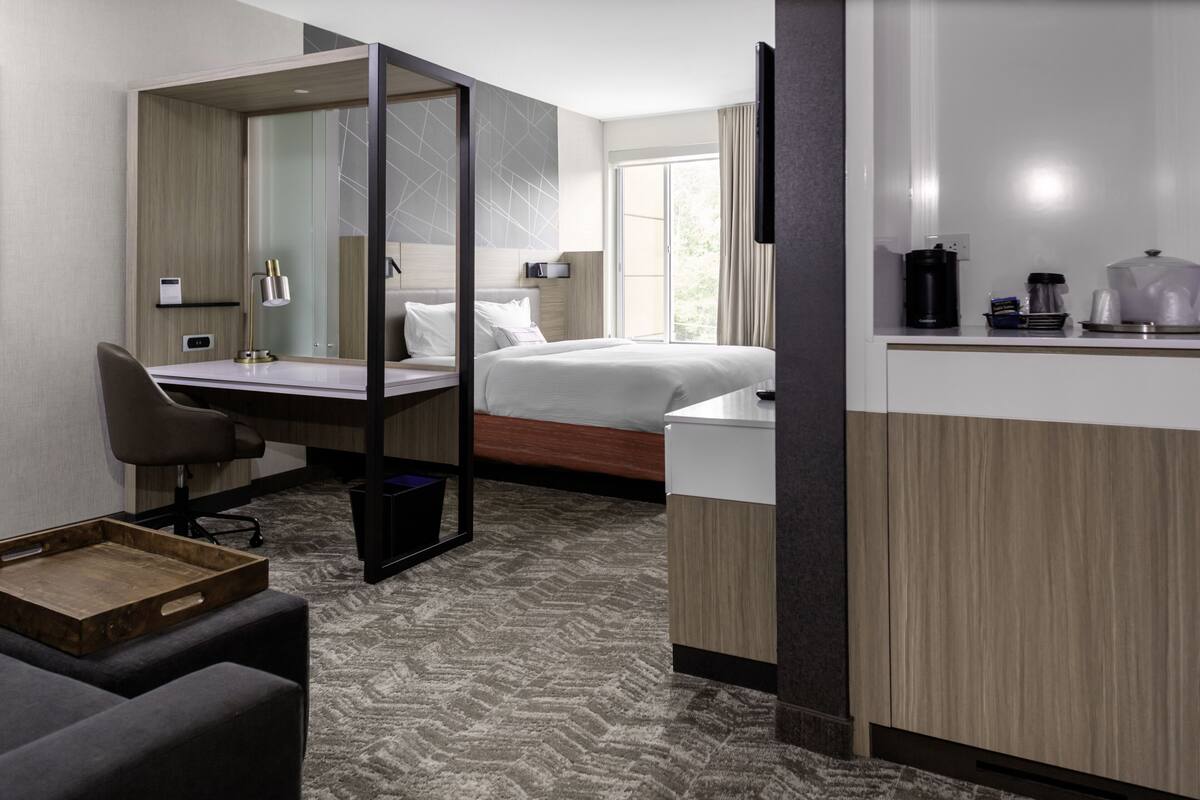 A modern hotel room with a bed, desk, chair, and kitchenette.