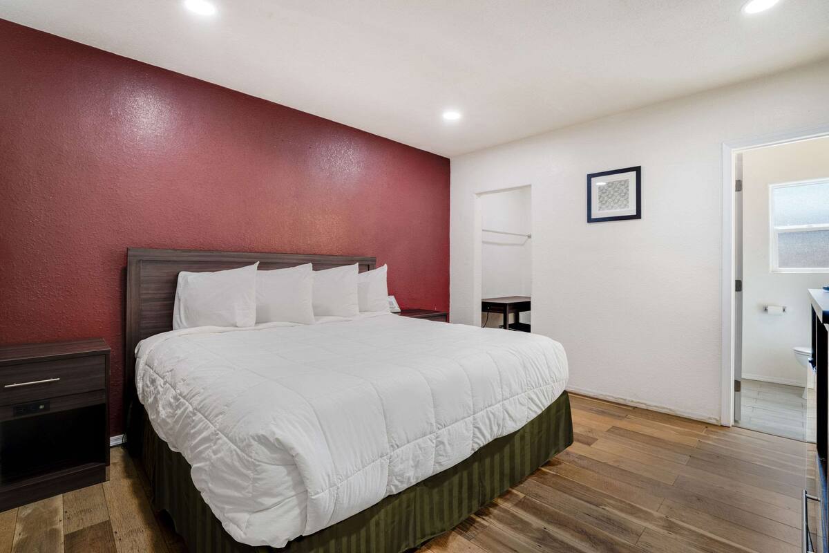 A bedroom with a large bed, a nightstand, a wooden floor, and a red accent wall.