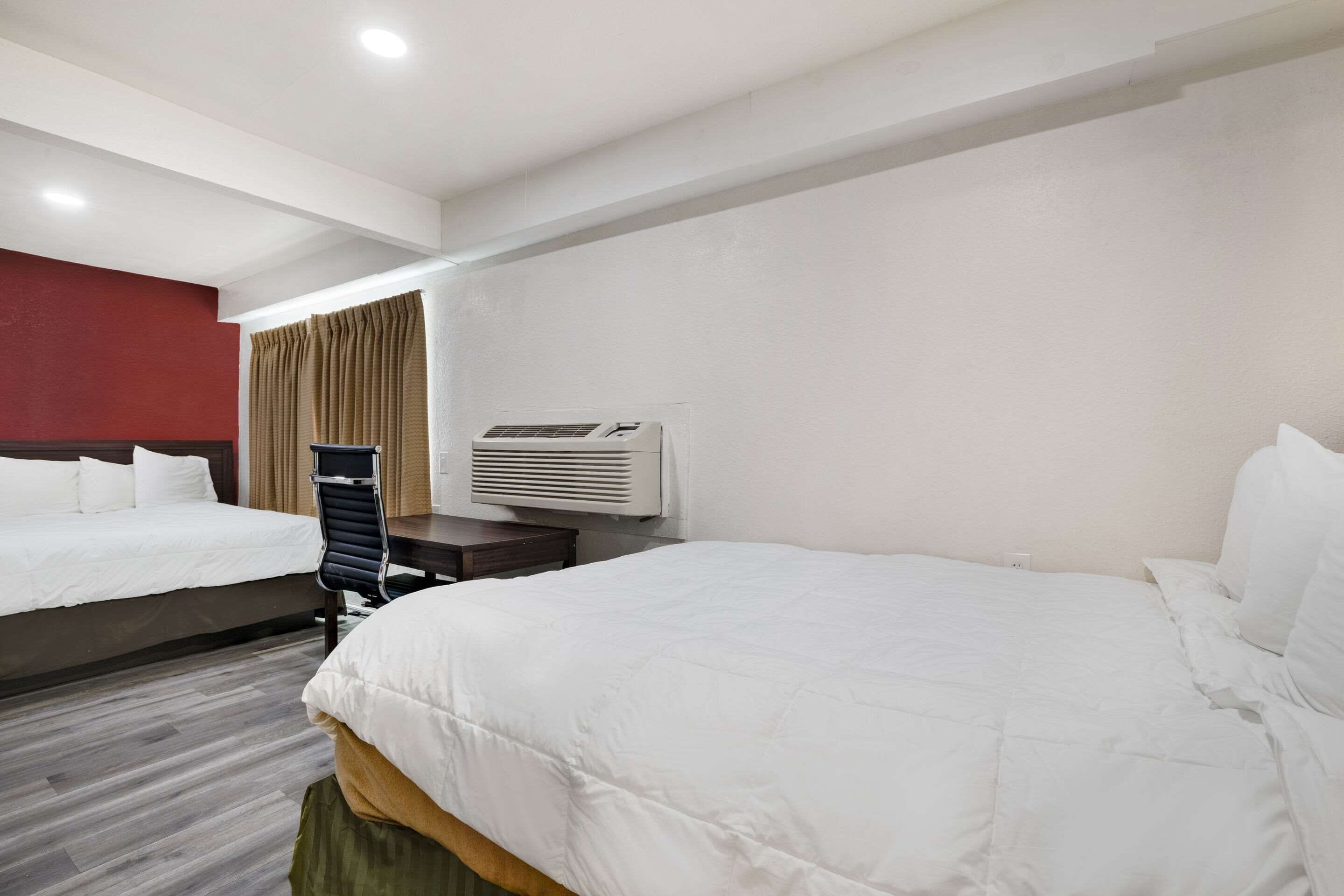 A hotel room with two beds, a desk, an air conditioning unit, and a window with curtains.