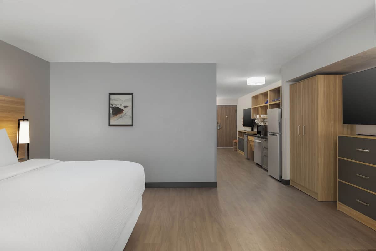 Hotelkamer met een bed, een bedlamp, een fotokader aan de muur, een kitchenette met kastjes en een televisie.