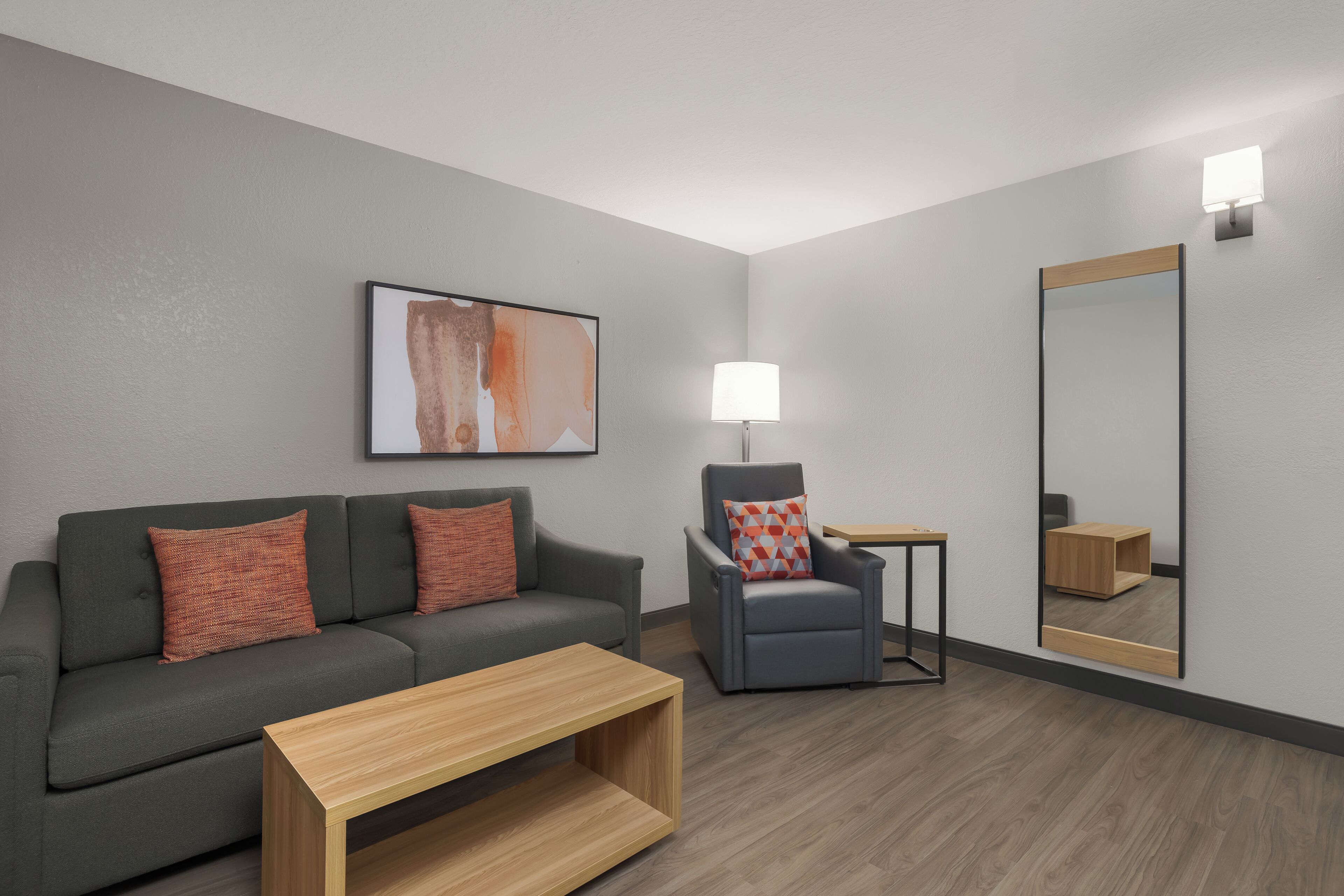 Una habitación moderna con un sofá gris, una mesita pequeña, una silla, una lámpara y un espejo grande.