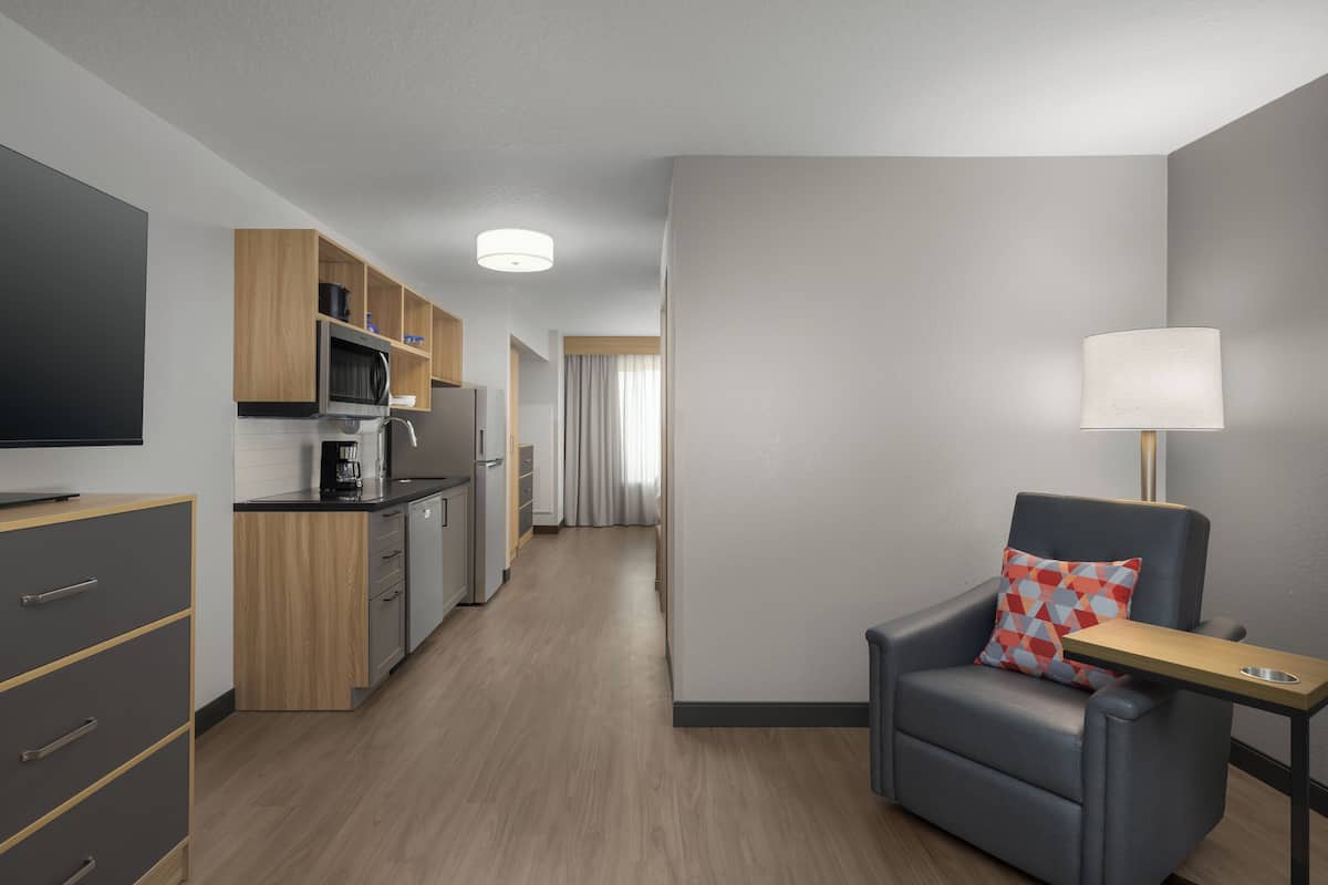 Een moderne hotelkamer met kitchenette, een flatscreen televisie, een comfortabele fauteuil met dienblad en een lamp.