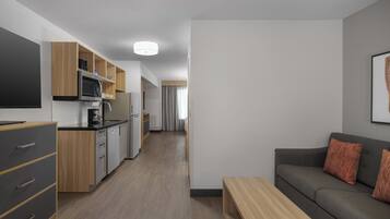 A modern kitchen with wooden cabinets, a microwave, and a coffee maker. There is a grey sofa with orange cushions and a wooden coffee table.