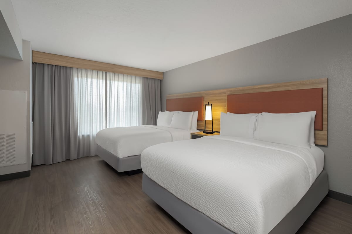 Een hotelkamer met twee bedden, een groot raam met gordijnen, een houten hoofdeinde en een nachtkastje met een lamp.
