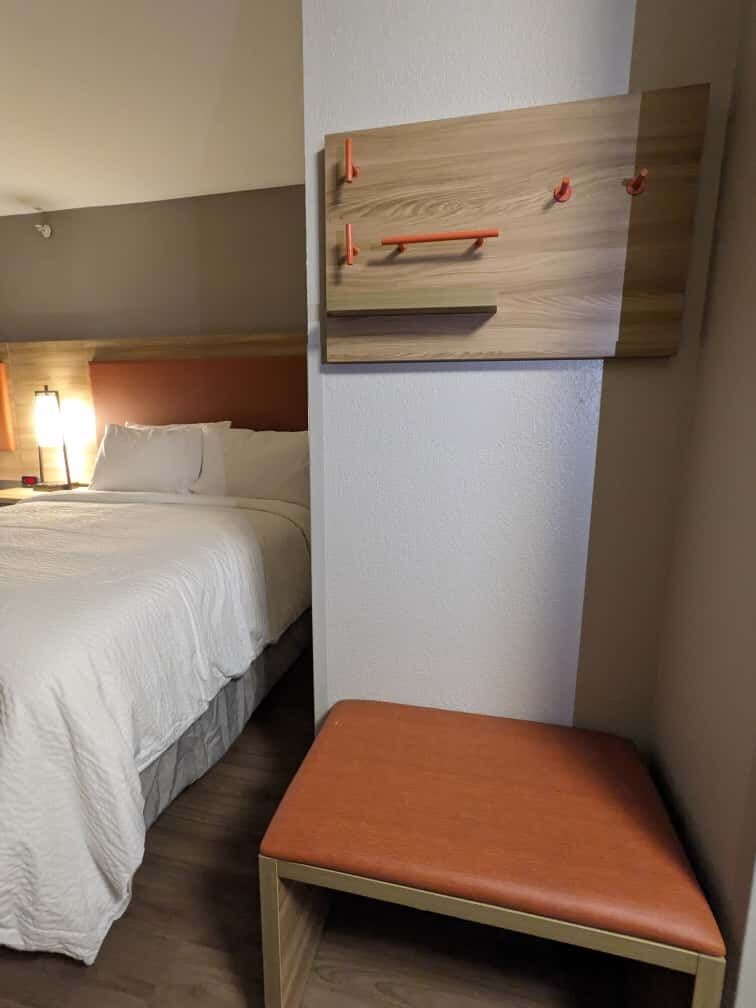 Ein Hotelzimmer mit Bett, Nachttisch mit Lampe und einer Holzwand mit Haken.