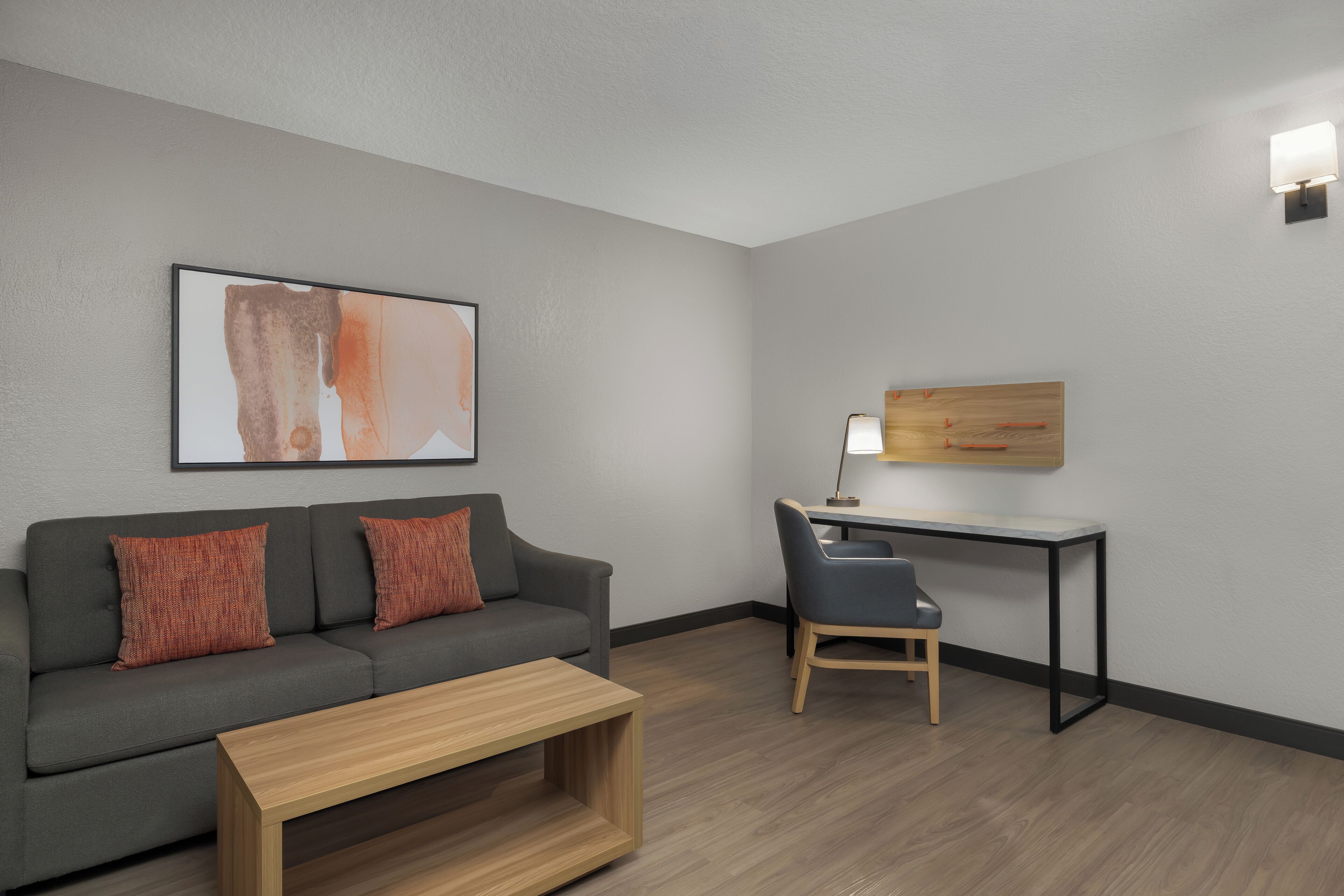 Een moderne kamer met een grijze bank, een houten salontafel, een bureau met stoel, en een wandlamp.