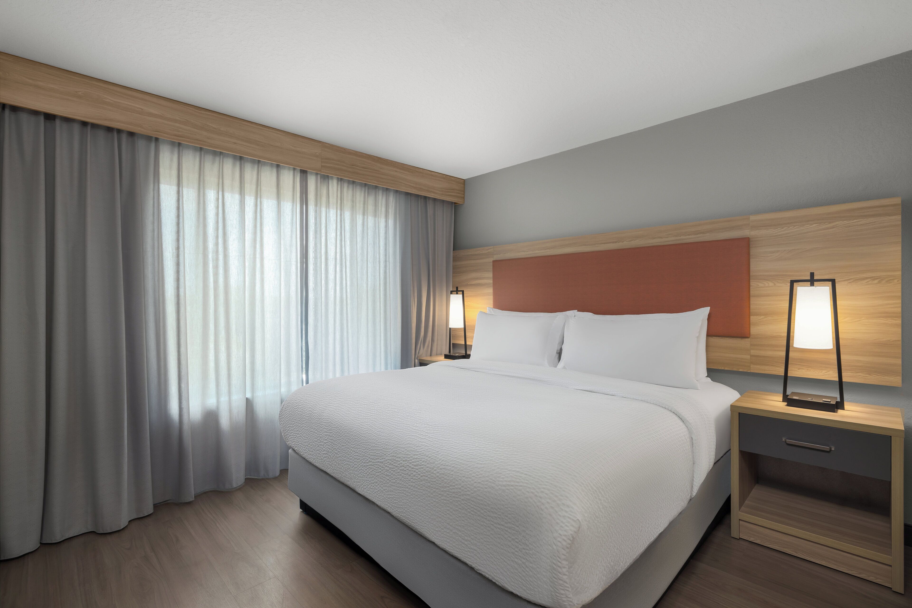 A hotel room with a large bed, wooden headboard, two bedside lamps, a nightstand, and a window with curtains.