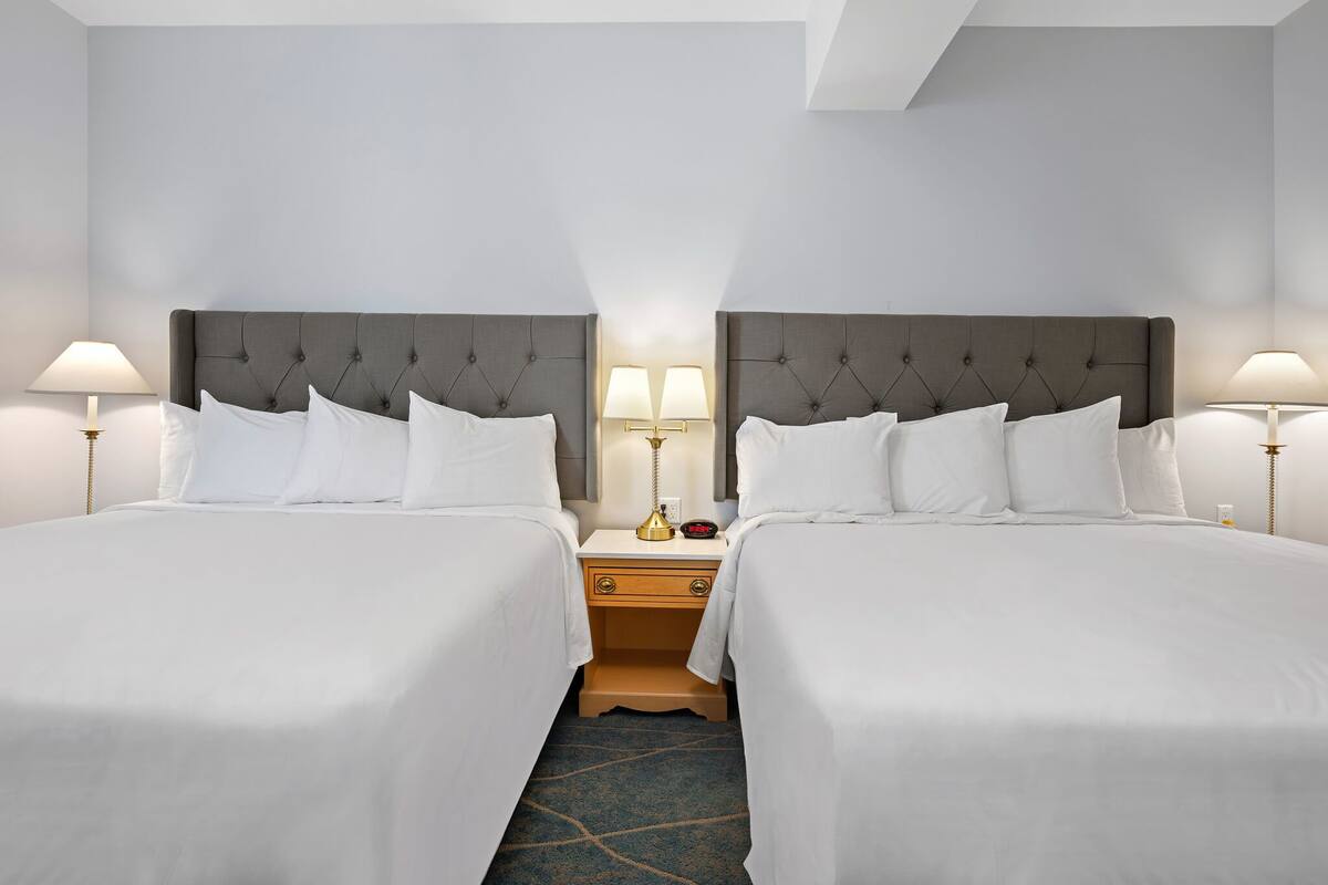 Ett hotellrum med två sängar, var och en med en grå klädd sänggavel, vita sängkläder och sänglampor.