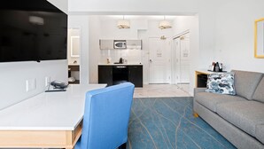 Una habitación de hotel moderna con una mesa de comedor blanca, una silla azul, un sofá gris y una cocina al fondo.