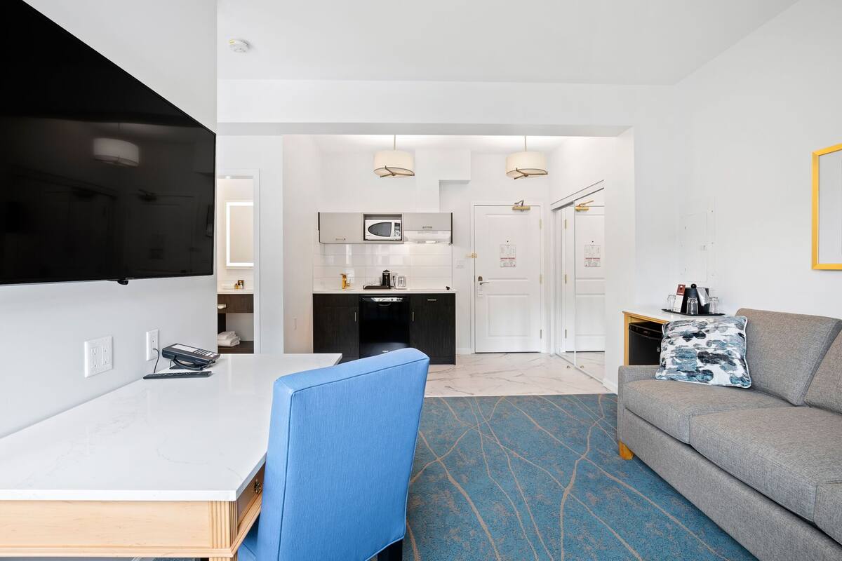 Ett modernt hotellrum med ett vitt matbord, en blå stol, en grå soffa och ett köksområde i bakgrunden.
