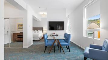 Una habitación de hotel con una mesa de comedor de vidrio, sillas tapizadas en azul, una cama con ropa blanca, un ventanal con vistas y un cuadro en la pared.