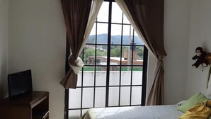 Une chambre d’hôtel avec un lit, un téléviseur posé sur un meuble, une fenêtre avec des rideaux et une vue sur les bâtiments et les collines environnantes.