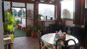 Ein überdachter Patio-Bereich mit einem runden Tisch, Stühlen und einer Couch. Es gibt Blumentöpfe, eine Backsteinmauer und einen Blick auf Gebäude im Hintergrund.