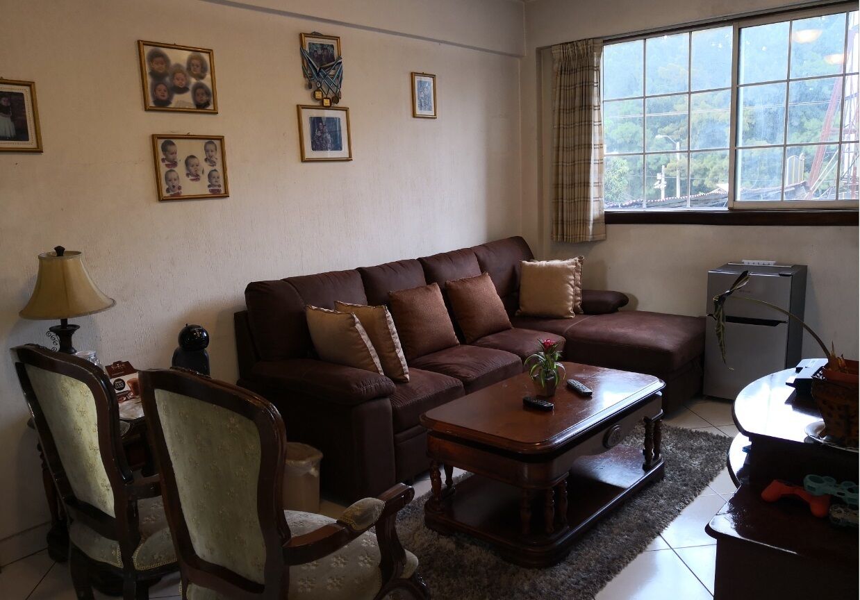 Una sala de estar con un sofá marrón, una mesa de centro de madera y una lámpara. En la pared hay cuadros enmarcados y una ventana con vistas a zonas verdes.