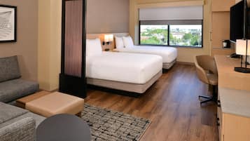 Quarto de hotel com duas camas, uma escrivaninha, uma cadeira, um sofá e uma janela grande com vista para a cidade.