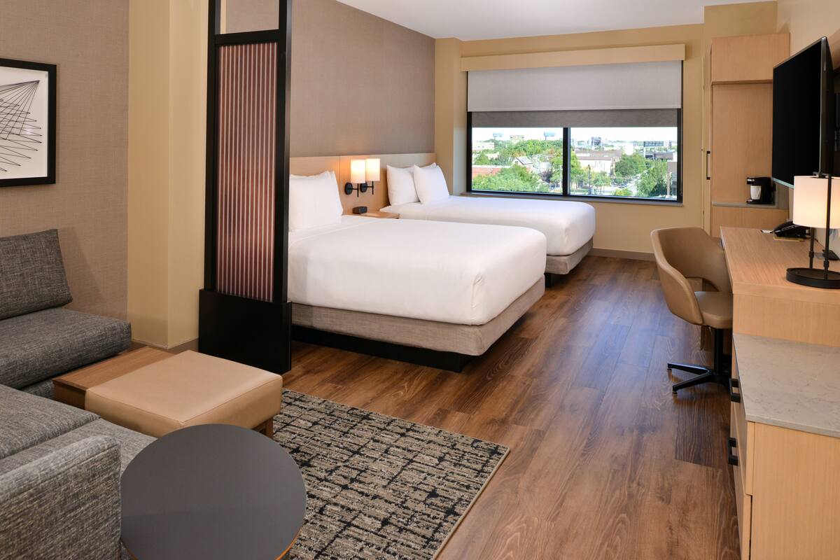 A hotel room with two beds, a sofa, a desk, and a large window offering a city view.