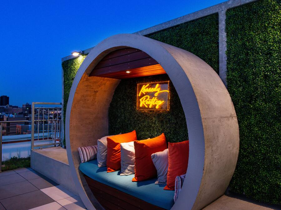 A modern outdoor seating area with a neon sign, surrounded by greenery and overlooking a cityscape.