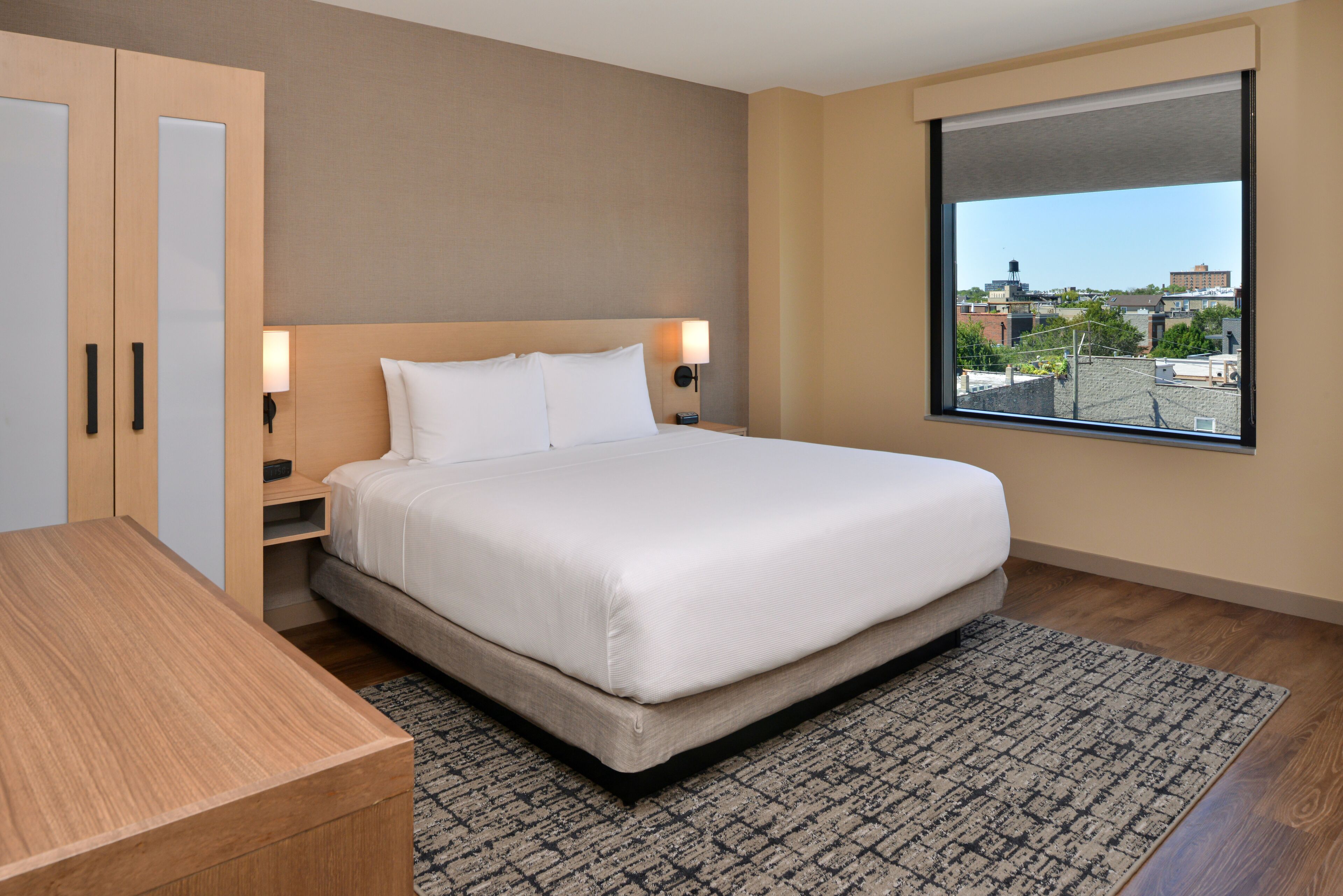 Une chambre d’hôtel moderne avec un grand lit, une armoire en bois, des tables de chevet et une vue sur la ville par une grande fenêtre.