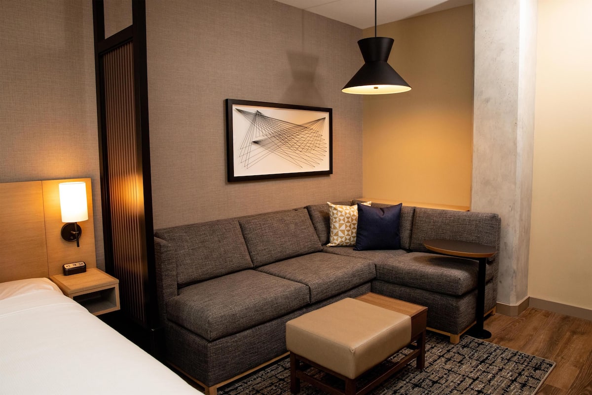 A modern hotel room with a sectional sofa, a small table, a bedside table with a lamp, and a framed abstract artwork on the wall.