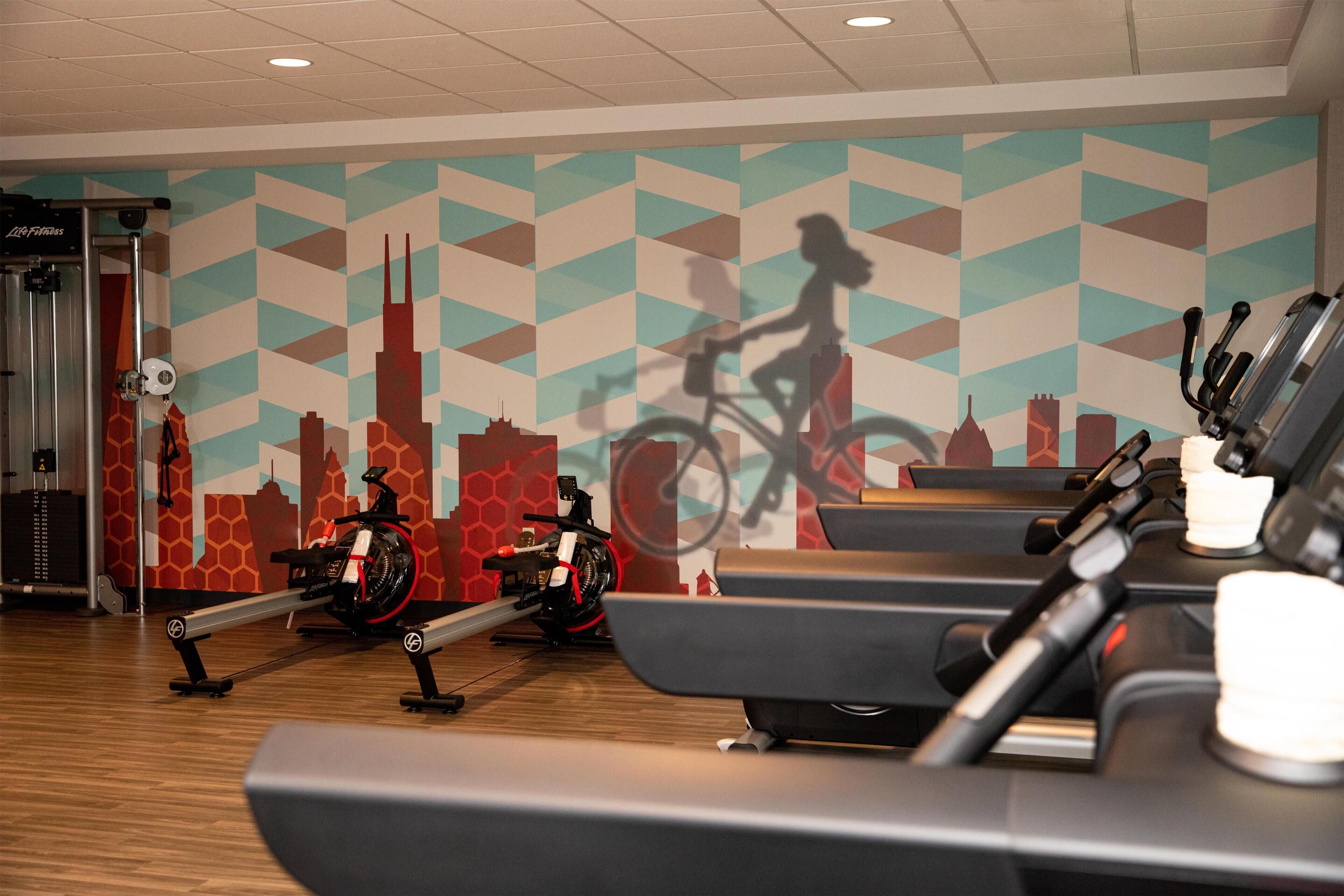 A gym with stationary bikes, treadmills, and a cityscape mural on the wall.