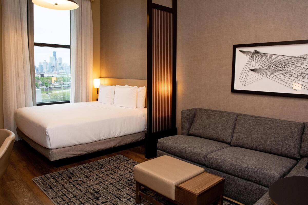 A hotel room with a bed, a sofa, a small table, a chair, and a city view through the window.