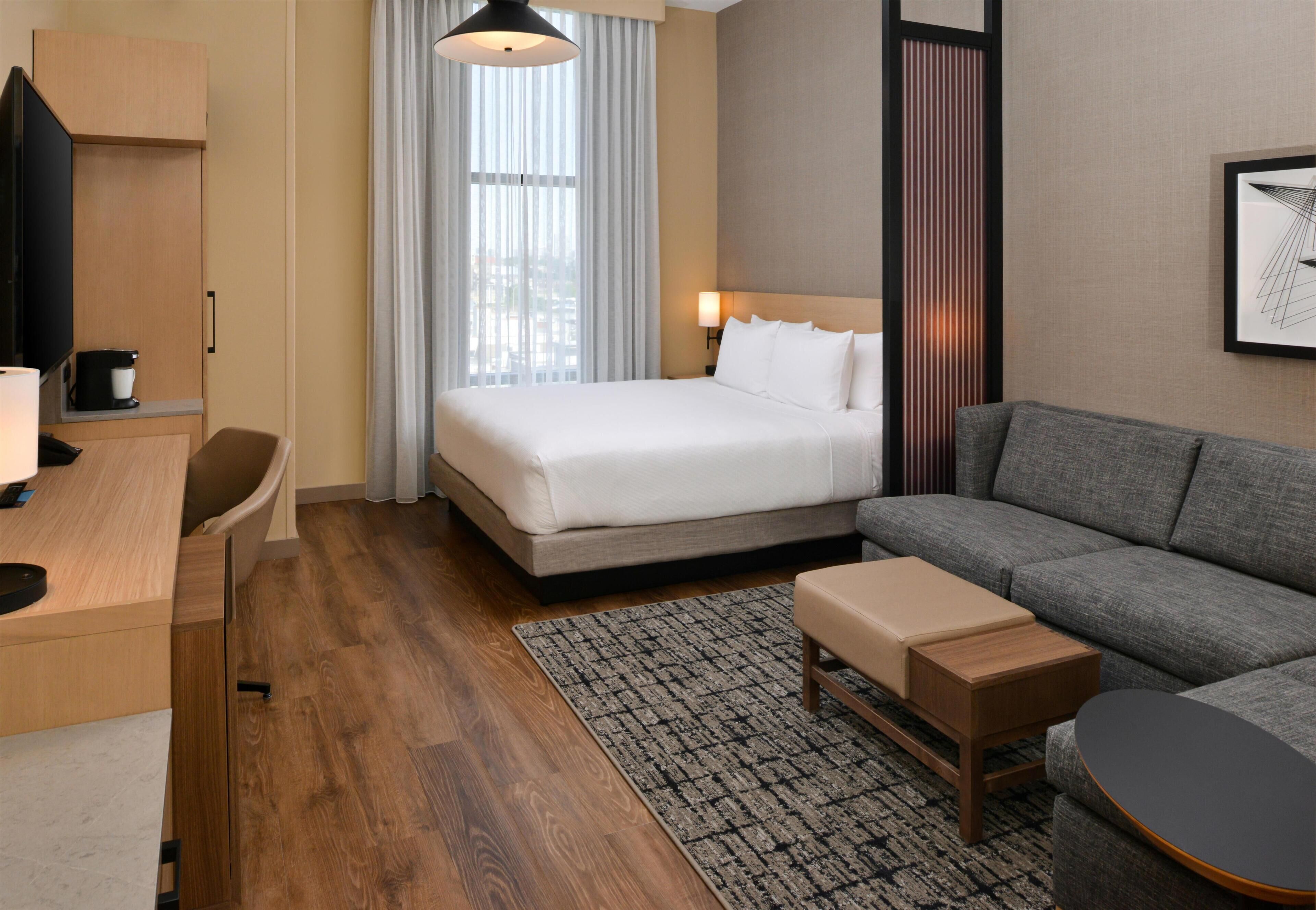 A hotel room with a bed, a sofa, a desk, a TV, and a window with curtains.