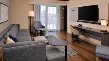 A modern hotel room with a sofa set, a small round table, a desk with a chair, a TV mounted on the wall, and a large window with curtains.