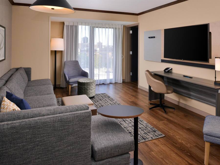 A modern hotel room with a sofa set, a small round table, a desk with a chair, a TV mounted on the wall, and a large window with curtains.