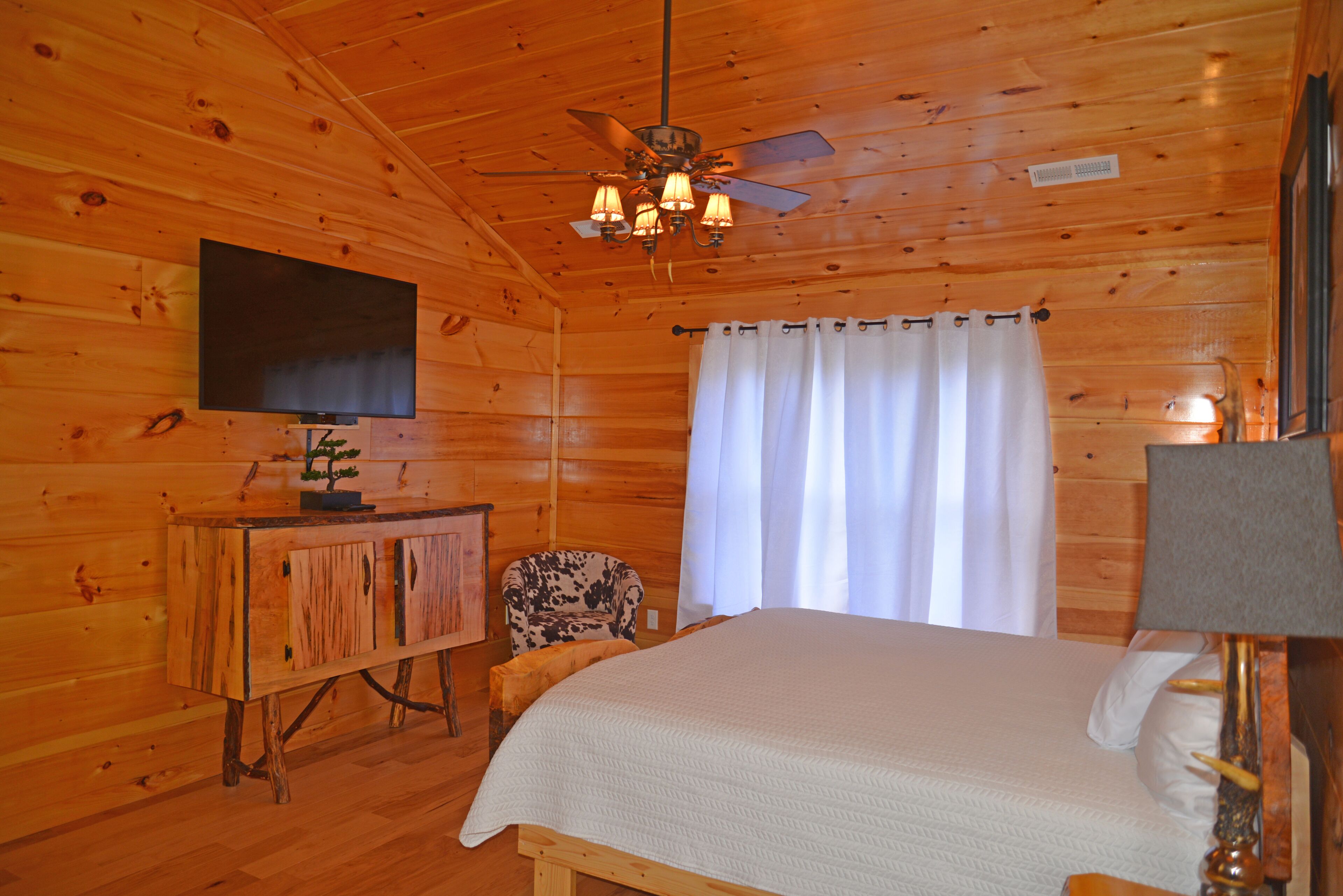 A wooden cabin room with a bed, a television on a wooden cabinet, a ceiling fan, and a window with curtains.