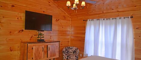 A wooden cabin room with a bed, a television mounted on the wall, a ceiling fan, and a small cabinet with a plant.