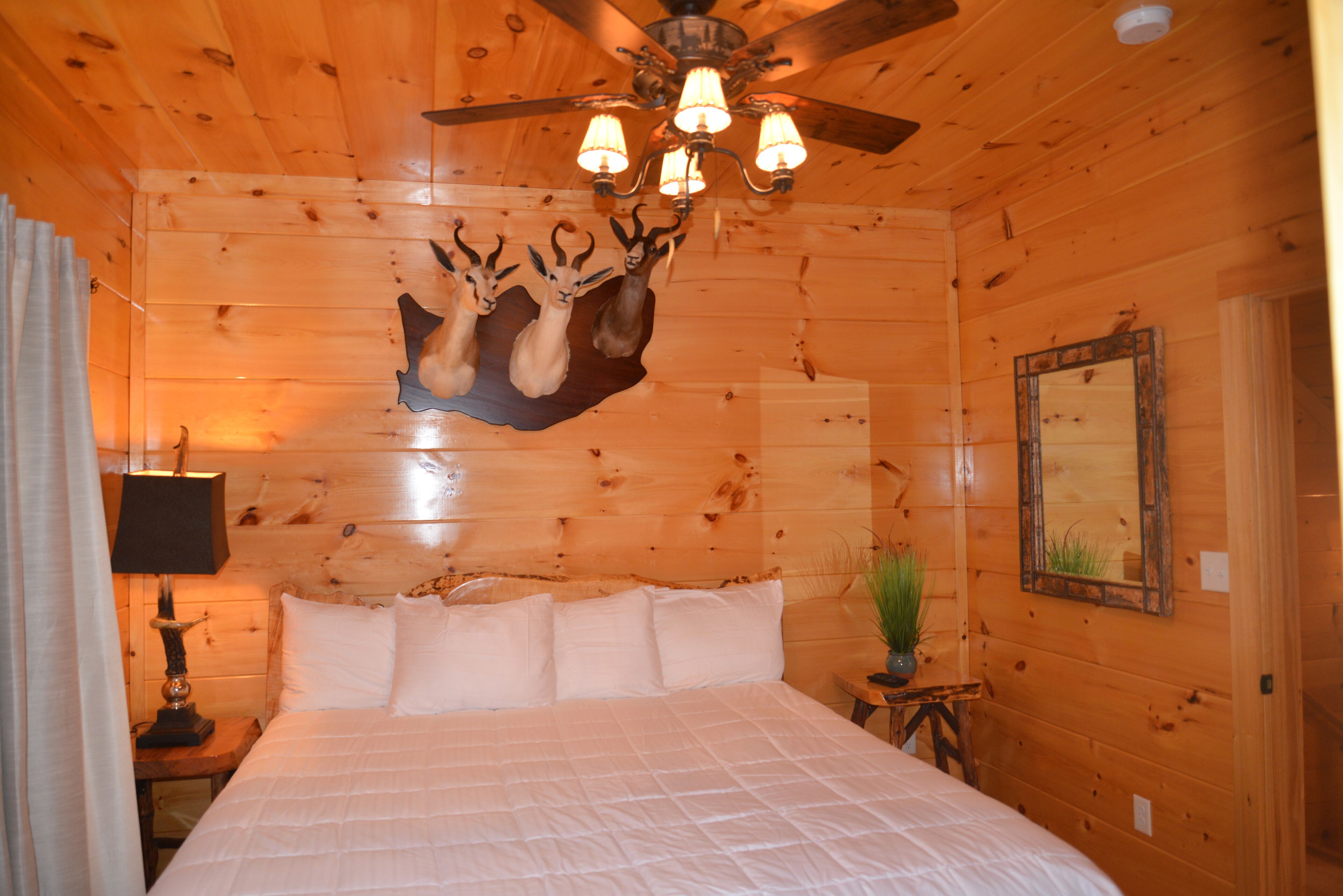 Ein Schlafzimmer in einer Holzhütte mit einem Bett, einem Deckenventilator, einem aufgehängten Hirschgeweih, einer Lampe und einem Spiegel.