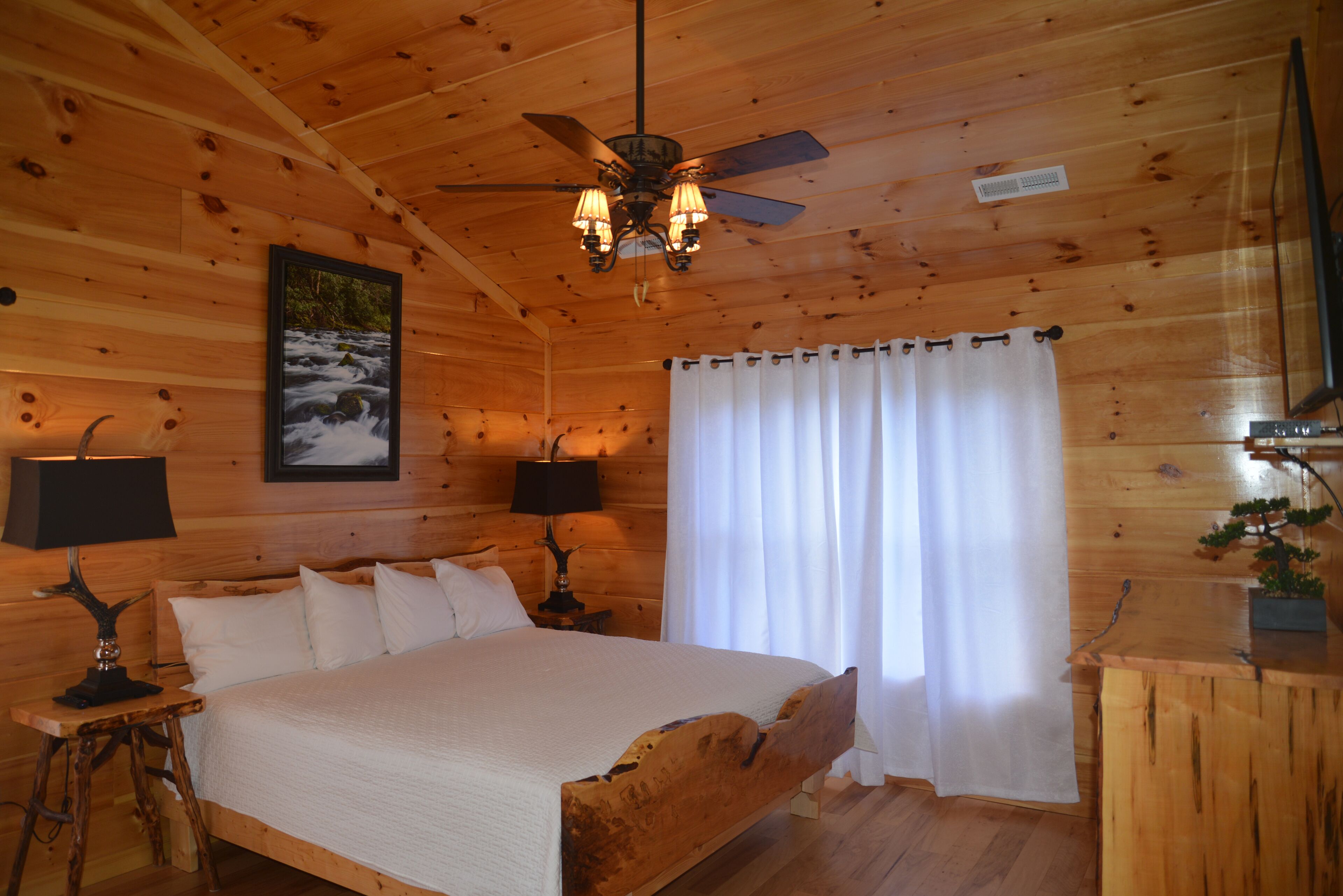 Un dormitorio de cabaña de madera con una cama, dos mesitas de noche con lámparas, un ventilador de techo, una ventana con cortinas y un cuadro en la pared.