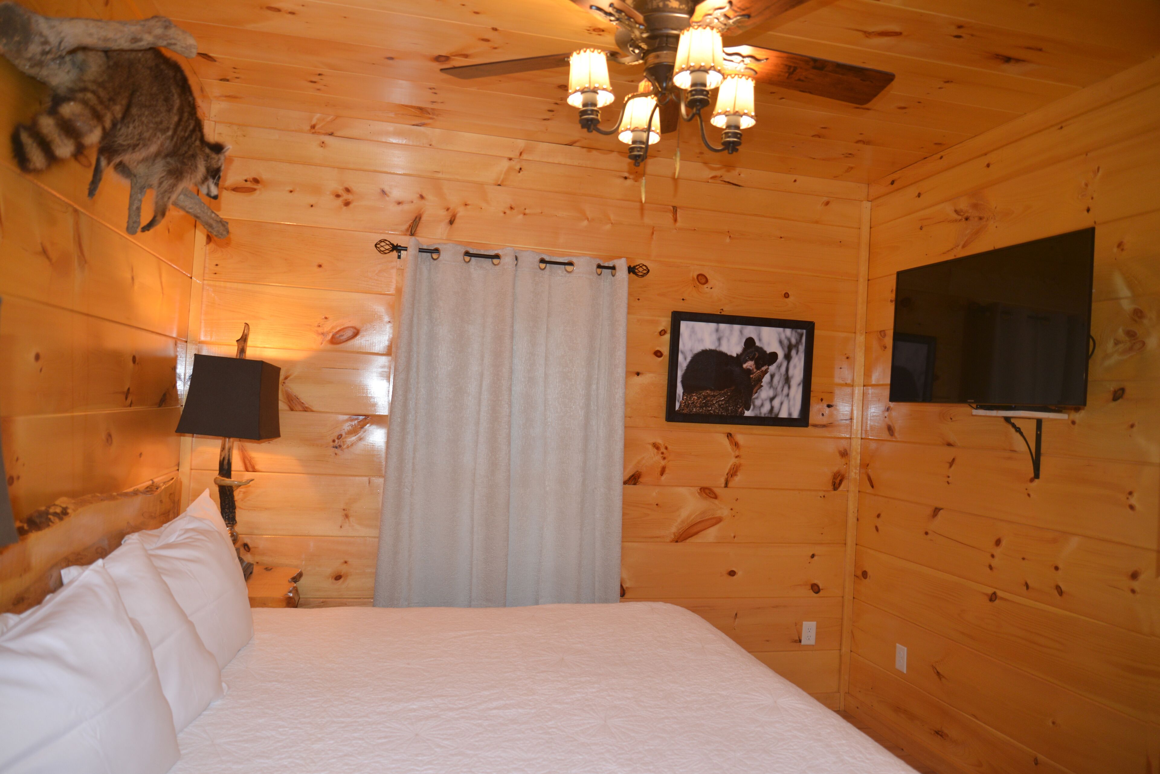 Quarto de cabana de madeira com cama, ventoinha no teto, televisão montada e luminária na parede.