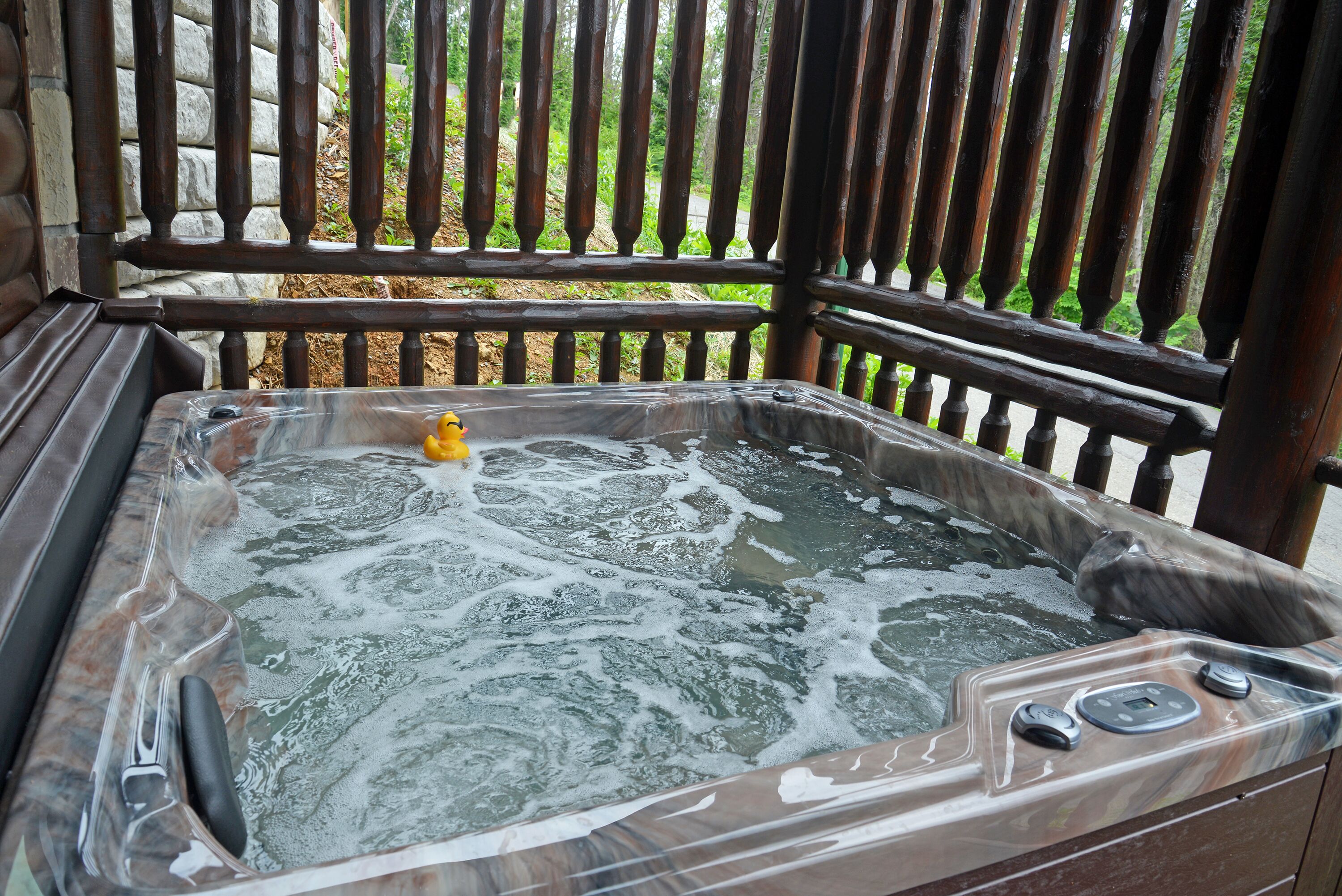 Un jacuzzi con un patito amarillo, rodeado de barandillas de madera y un muro de piedra.