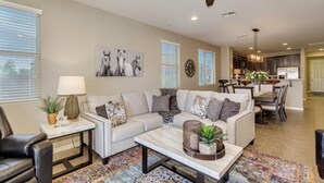 A modern living room with a sofa, coffee table, and decorative pillows. There is a dining area with a table and chairs, and a kitchen in the background. - Modern home in Estrella Mtn Ranch - refreshing Pool & Spa / Fire pit / Mtn Views (Goodyear)