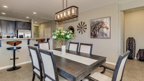 A dining area with a dark wooden table, white cushioned chairs, a vase with flowers, a wall-mounted light fixture, and a horse painting. - Modern home in Estrella Mtn Ranch - refreshing Pool & Spa / Fire pit / Mtn Views (Goodyear)