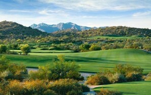 Um campo de golfe com colinas verdes ondulantes, árvores e montanhas ao fundo.