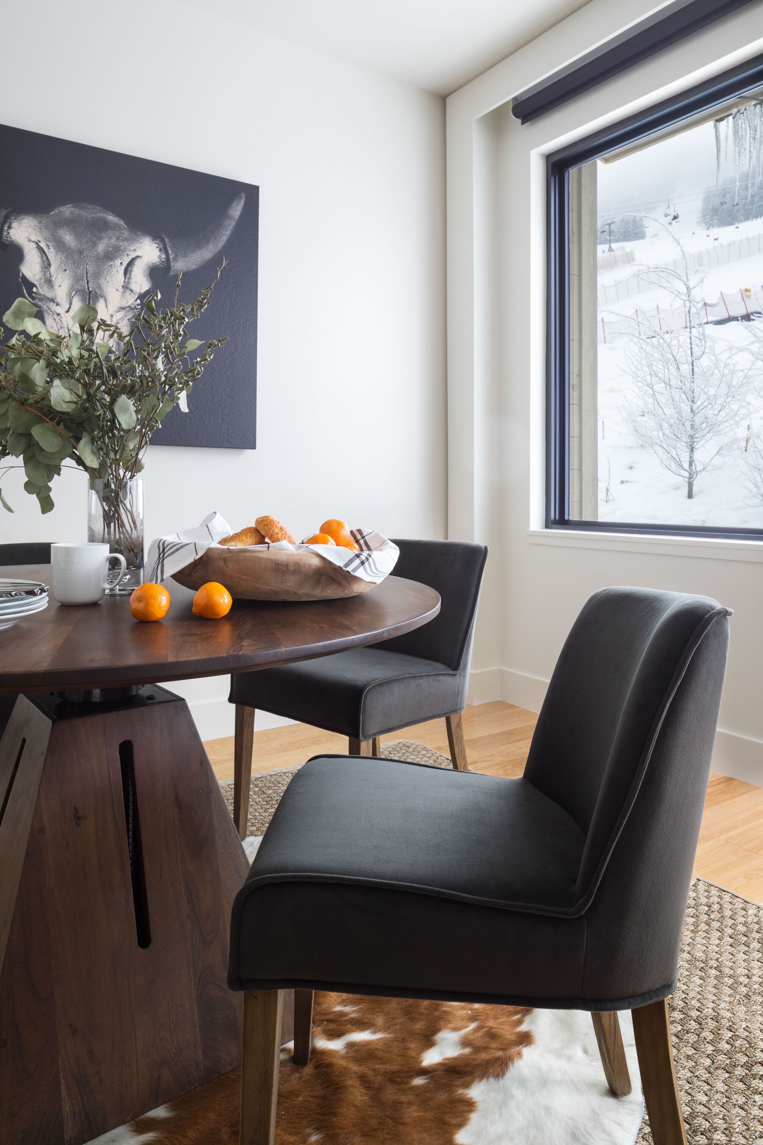 A modern dining area with a wooden table, black chairs, a vase with green leaves, a bowl of oranges, and a painting of a bull's head on the wall.