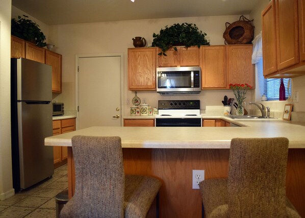A kitchen with wooden cabinets, a stainless steel refrigerator, a microwave, an oven, and a sink. There are two chairs and a small island with a countertop. - Lovely First Floor Condo in Small, Gated Community (Goodyear)