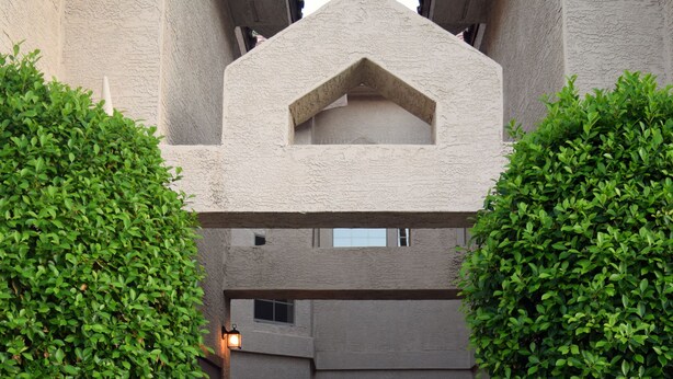 Une entrée d’immeuble avec un chemin bordé de deux arbres, une porte et une petite fenêtre située au-dessus de la porte.