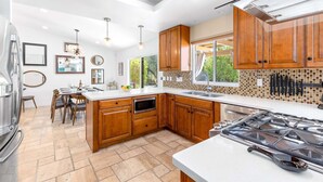 A modern kitchen with wooden cabinets, a gas stove, a dining area with a table and chairs, and a view of greenery outside. - Modern Loma Vista – Private Pool & Spa Retreat (Palm Springs)