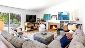 A living room with a sofa, ottoman, TV, fireplace, bookshelves, and a view through a glass door. - Modern Loma Vista – Private Pool & Spa Retreat (Palm Springs)