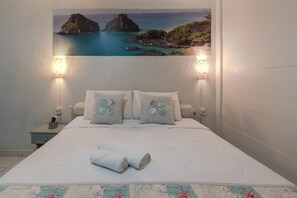 A neatly made bed with floral-patterned pillows and a rolled towel, a nightstand with a lamp, and a wall-mounted picture of a coastal landscape. - Pousada Carmo (Fernando de Noronha)