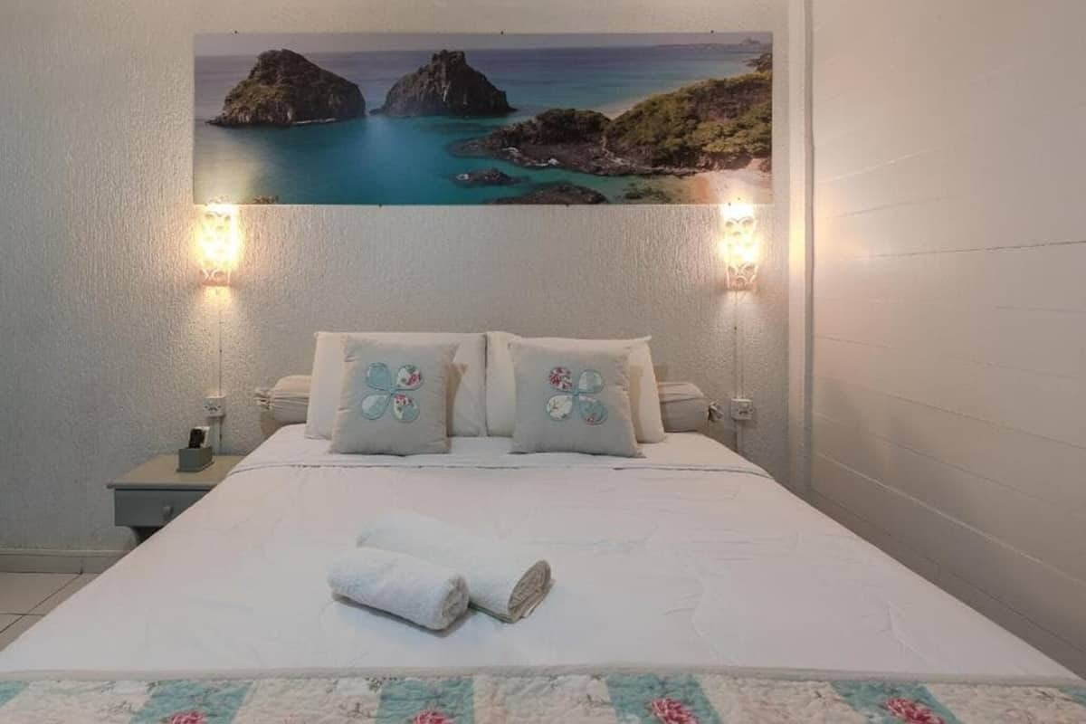 A bedroom with a bed, pillows, a nightstand, a wall-mounted picture, and a wall-mounted light.