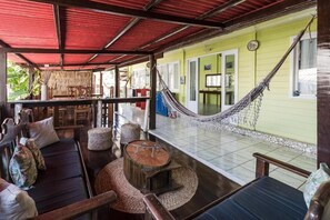 A covered outdoor seating area with a hammock, wooden furniture, and a green building in the background. - Pousada Carmo (Fernando de Noronha)