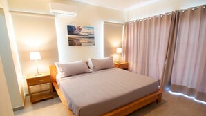 Une chambre à coucher avec un lit en bois, deux tables de chevet avec des lampes, une petite table, un climatiseur fixé au mur et une fenêtre avec des rideaux.
