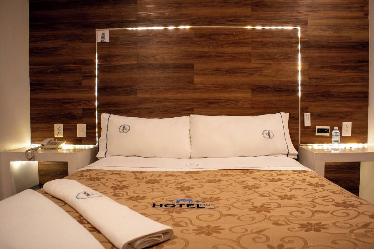 A hotel room with a wooden headboard, a bed with white linens, a patterned bedspread, a rolled towel on the bed, and a bedside table with a phone and a bottle of water.
