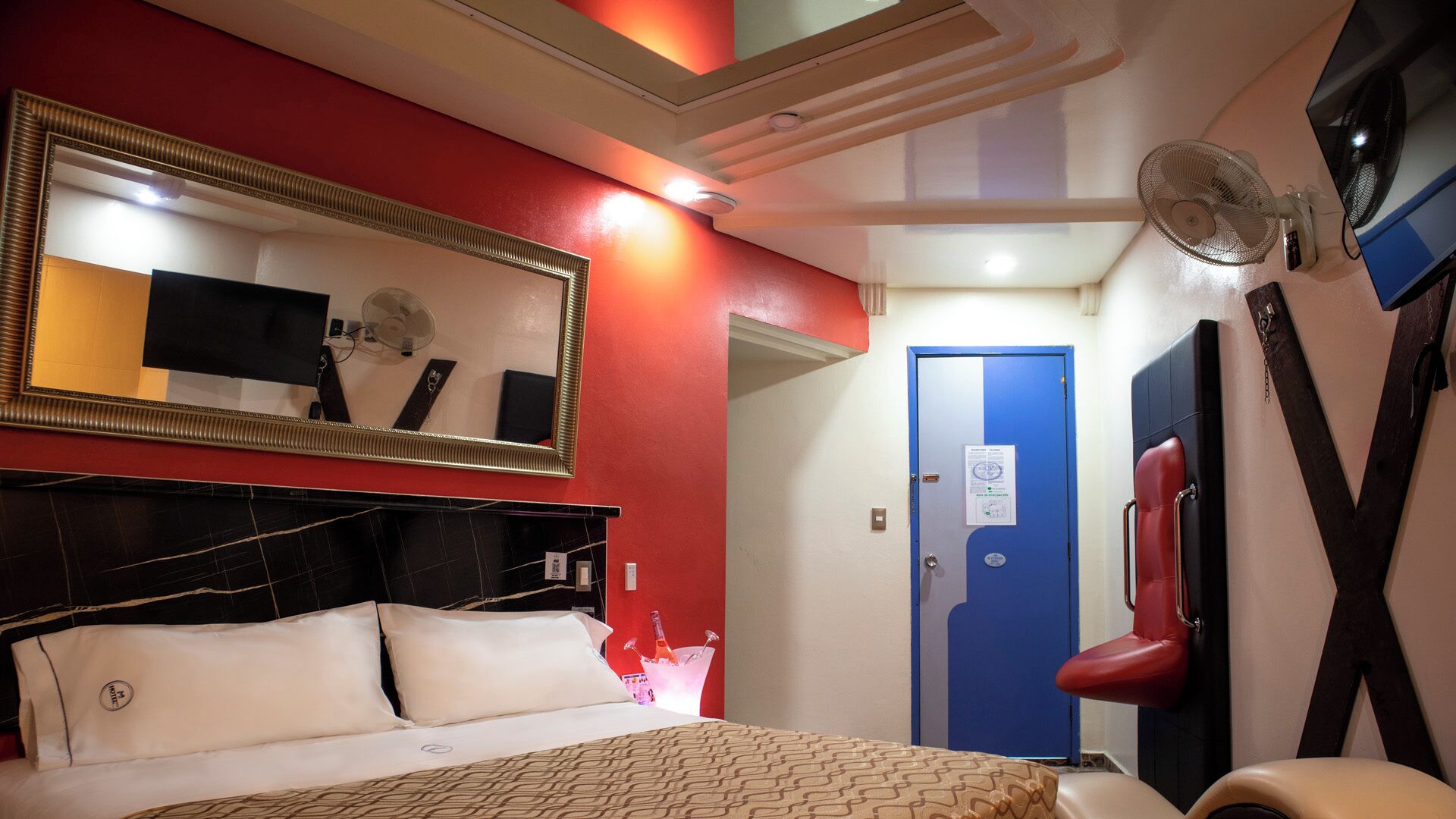 A hotel room with a bed, a mirror, a red wall, a blue door, and a fan.
